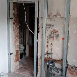 Stelaż z profili metalowych do płyty karton gips, widoczne fragmenty instalacji hydraulicznej, ślady po starej instalacji na ścianie, gruz i wiadro na podłodze.