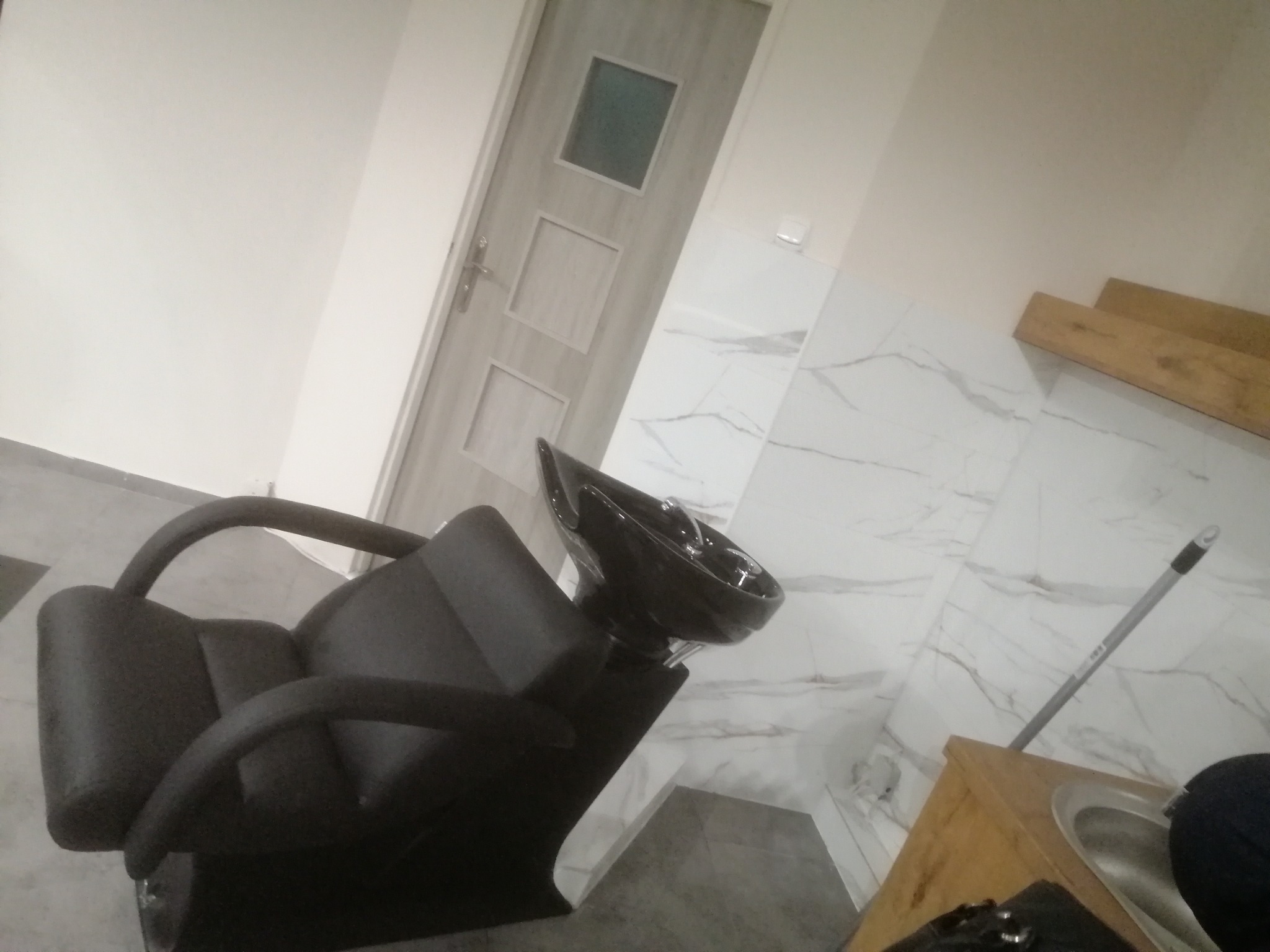 Czarne krzesło fryzjerskie z umywalką w łazience wykończonej marmurowymi płytkami, widoczny fragment drewnianej szafki i mop oparty o ścianę.