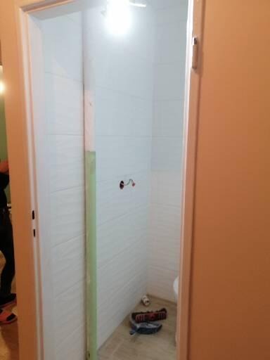 Wnętrze łazienki w trakcie remontu, widoczne świeżo położone białe płytki, narzędzia na podłodze i wystające kable. Drzwi wejściowe częściowo zasłaniają widok.