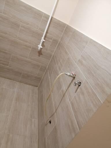 Płytki imitujące drewno na ścianie łazienki, widoczny biały rurociąg i wężyk podłączeniowy do baterii, niedokończona instalacja.