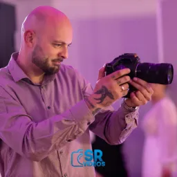 Łysy mężczyzna z brodą i tatuażami na rękach trzyma czarną kamerę filmową, skupiając wzrok na ekranie, w tle rozmyta postać i fioletowe oświetlenie.