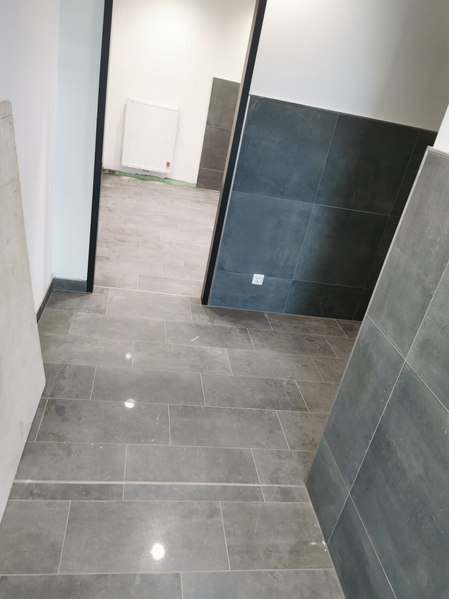 Szare, prostokątne płytki podłogowe i ścienne w trakcie montażu, widoczny fragment grzejnika i gniazdko elektryczne.