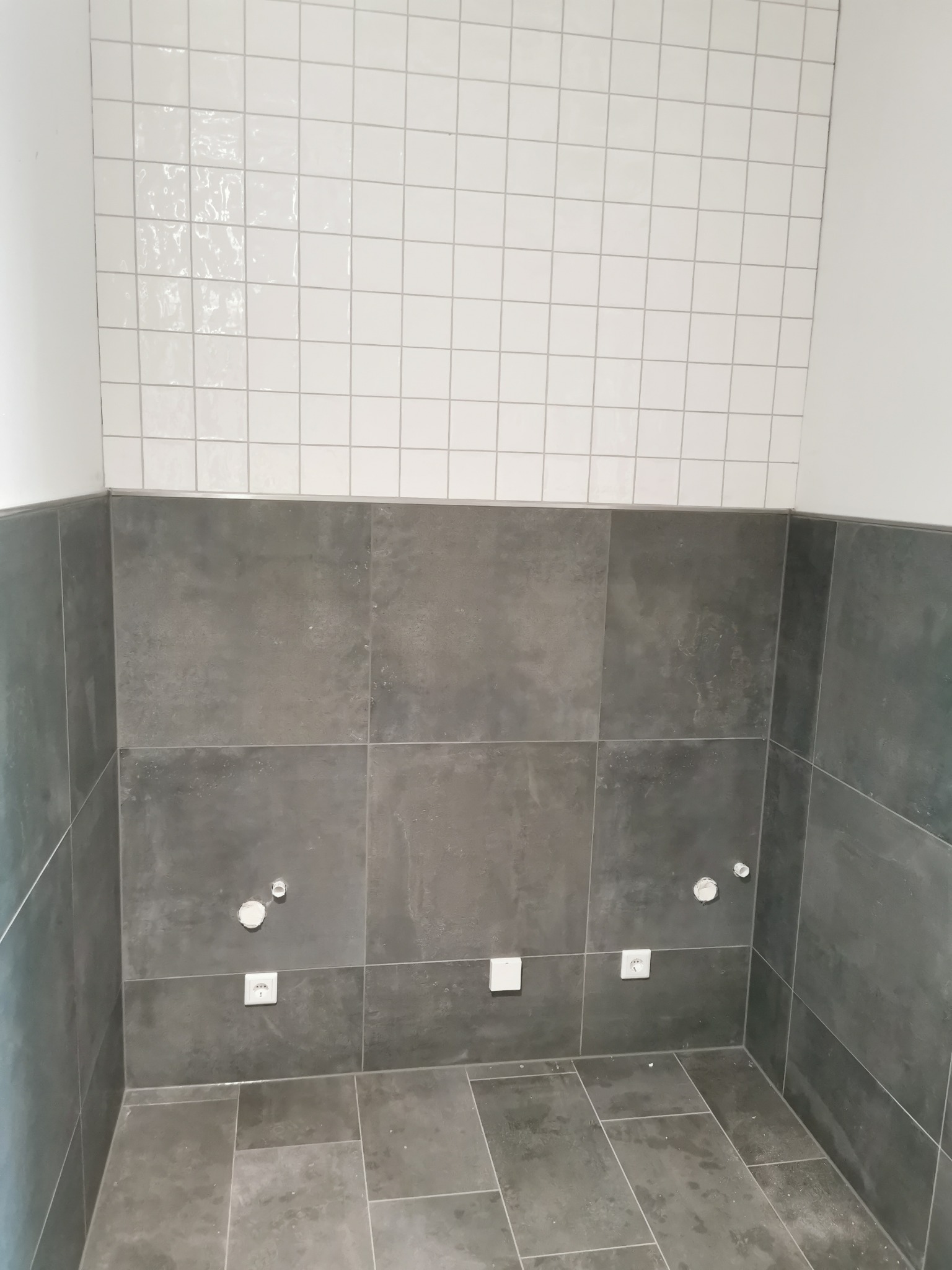 Ściana łazienki wyłożona częściowo białymi, błyszczącymi płytkami kwadratowymi, a częściowo szarymi, matowymi płytkami wielkoformatowymi. Widoczne gniazdka elektryczne i wyprowadzenia hydrauliczne.