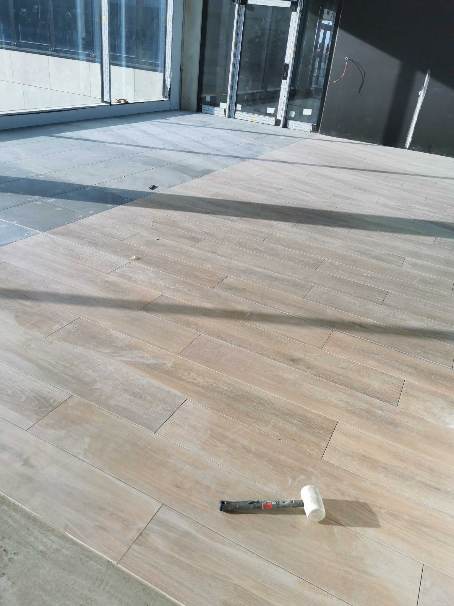 Układanie podłogi z płytek imitujących drewno, widoczny gumowy młotek glazurniczy i fragment podłogi z szarych płytek w surowym stanie.