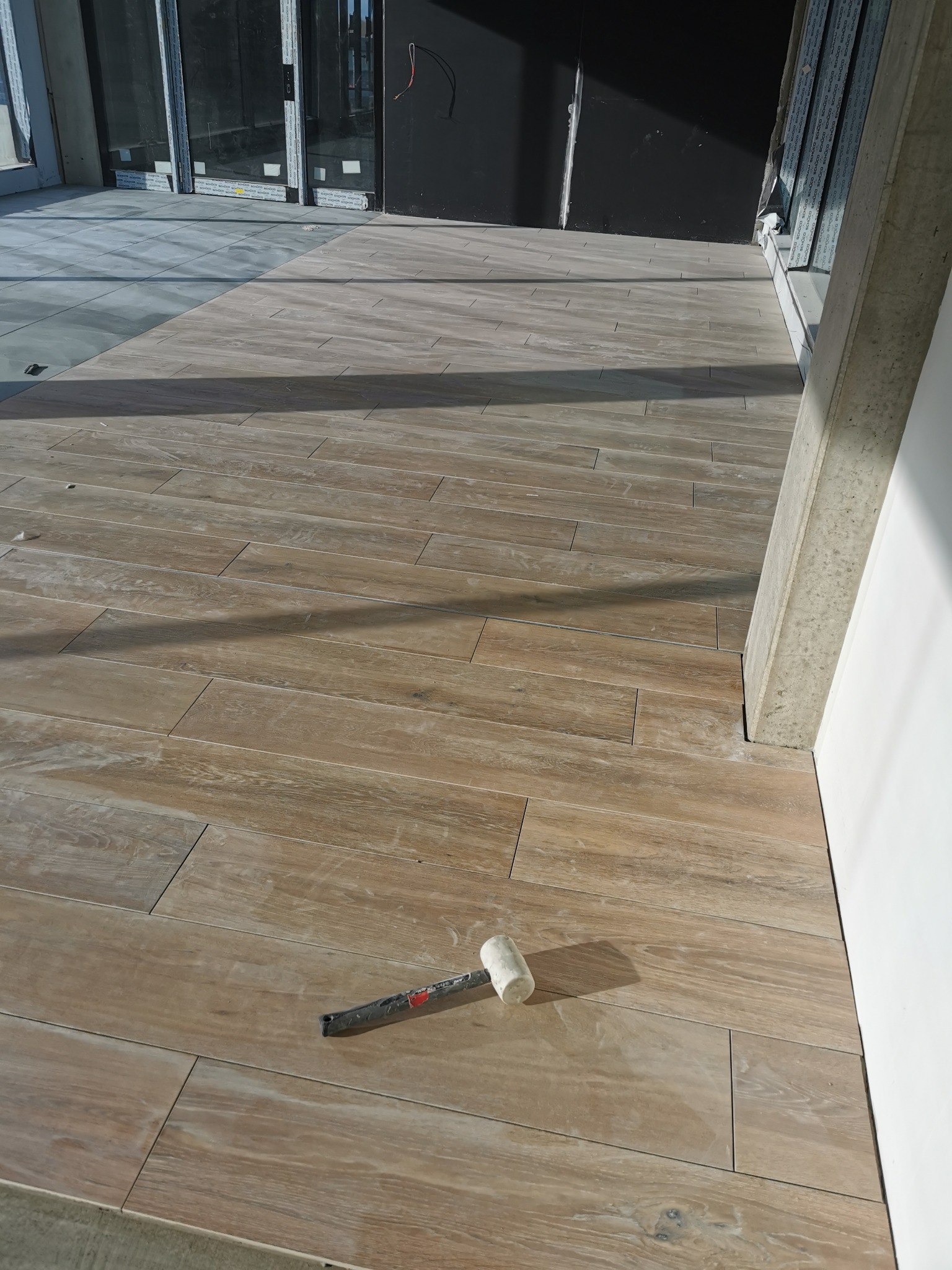 Układanie płytek podłogowych imitujących drewno w nowym budynku, widoczny gumowy młotek glazurniczy i fragmenty szarych płytek w sąsiednim pomieszczeniu.