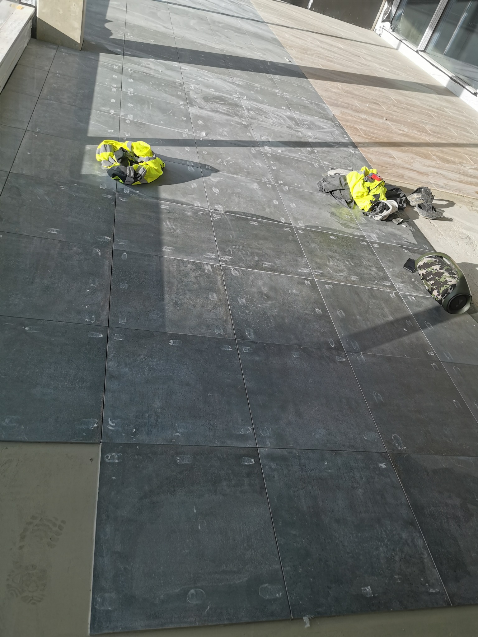 Ułożone, ciemne płytki podłogowe z widocznymi śladami fugowania, żółta kamizelka odblaskowa, buty robocze i głośnik przenośny na powierzchni.