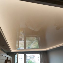 Lulux Krzysztof Cieślicki - Błyszczący, beżowy sufit napinany z oświetleniem LED wzdłuż obwodu, odbijający widok z okna na tle szarej ściany i częściowo widocznych mebli.