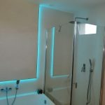 Nowoczesna łazienka z podświetleniem LED w kolorze turkusowym wokół wnęki prysznicowej i wanny, jasne płytki na ścianach, kabina prysznicowa ze szklanymi drzwiami.