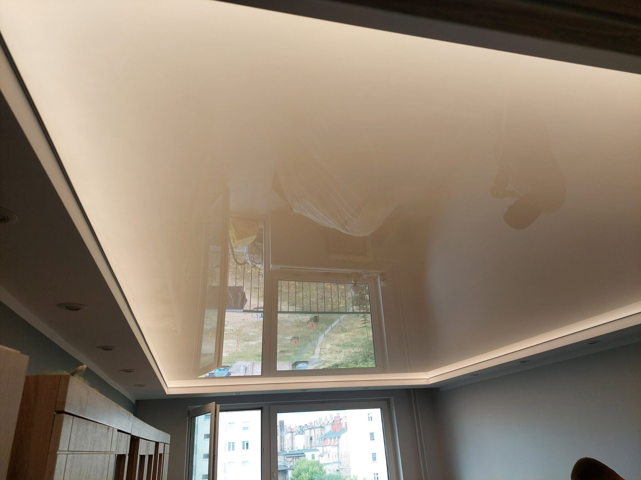 Błyszczący, beżowy sufit napinany z oświetleniem LED wzdłuż obwodu, odbijający widok z okna na tle szarej ściany i częściowo widocznych mebli.