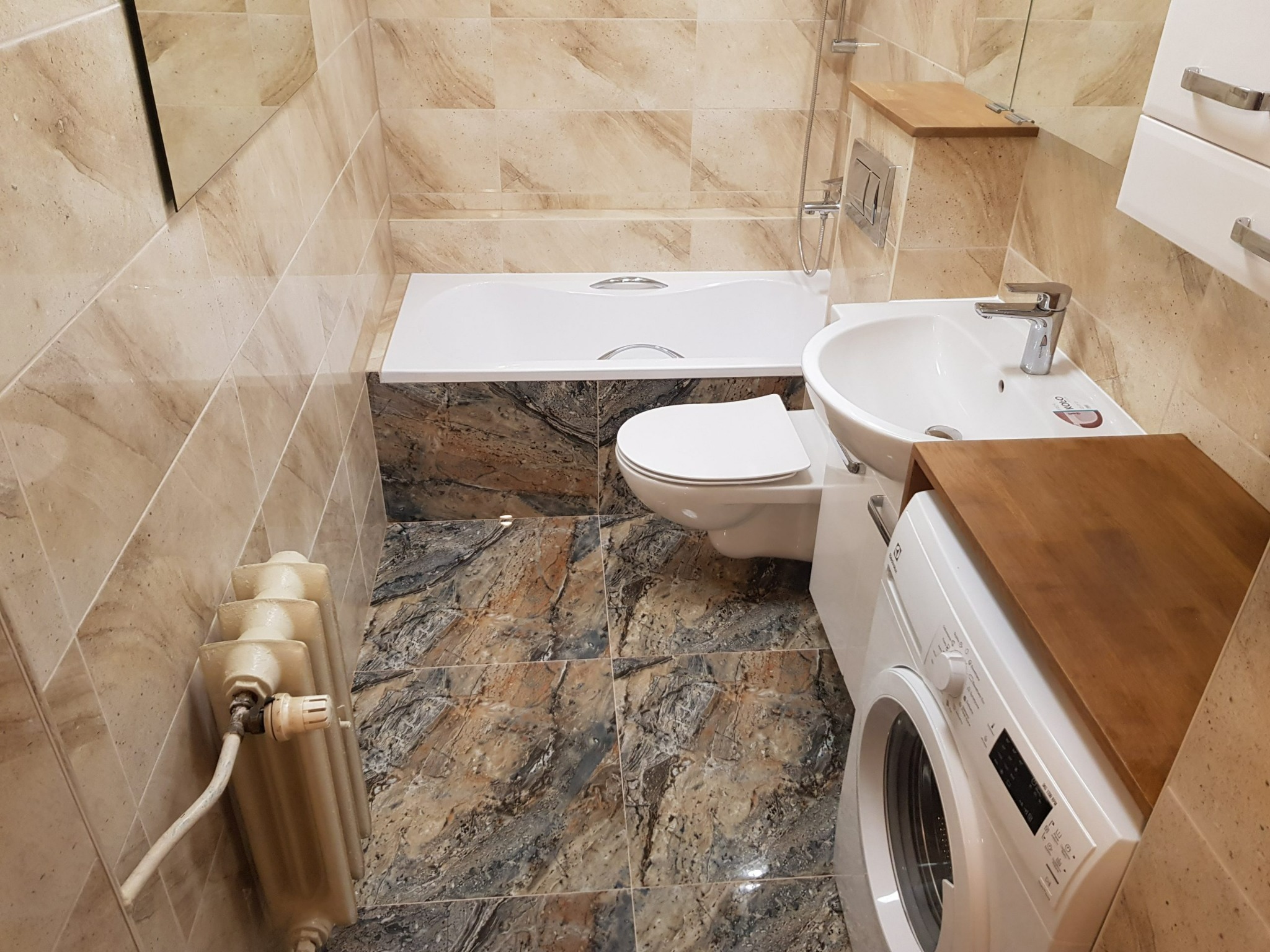 Ukończona łazienka z beżowymi płytkami imitującymi marmur, białą armaturą, grzejnikiem żeberkowym i pralką z drewnianym blatem.