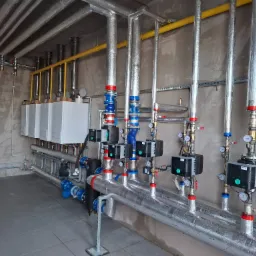 Kompleksowa instalacja grzewcza: kotły, rury w izolacji, zawory, manometry. Widoczne elementy instalacji gazowej i grzewczej w surowym wnętrzu.