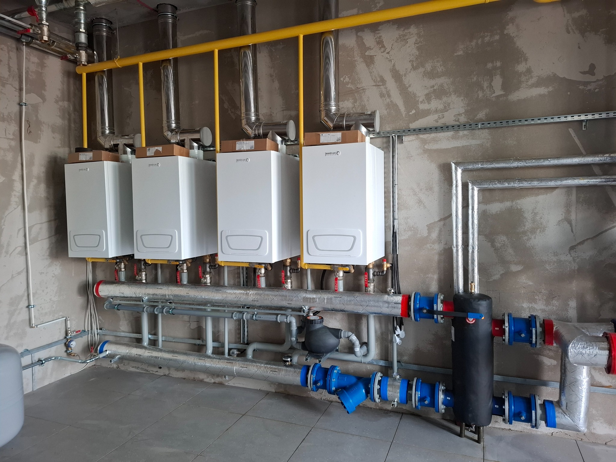 Cztery białe piece grzewcze z żółtą instalacją gazową i srebrnymi rurami na szarym tle ściany i podłogi. Instalacja połączona niebieskimi i czerwonymi złączkami.