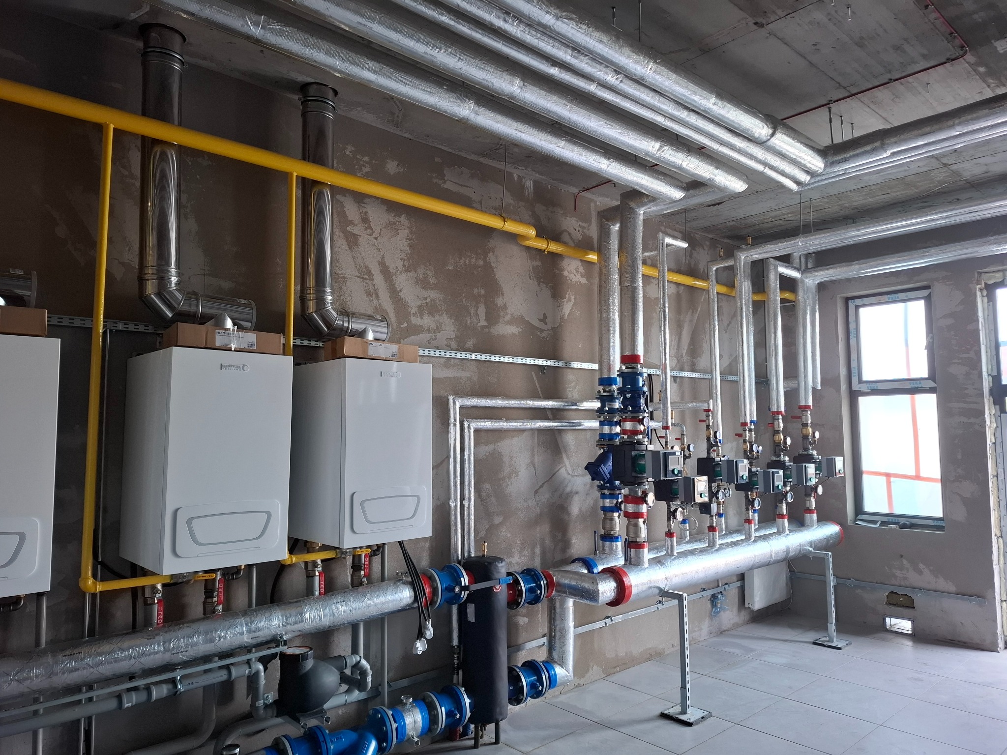 Kotłownia z trzema białymi piecami, żółtą rurą gazową i systemem rur grzewczych. Rury są zaizolowane, a ściany surowe, betonowe. Widoczne liczniki i zawory.