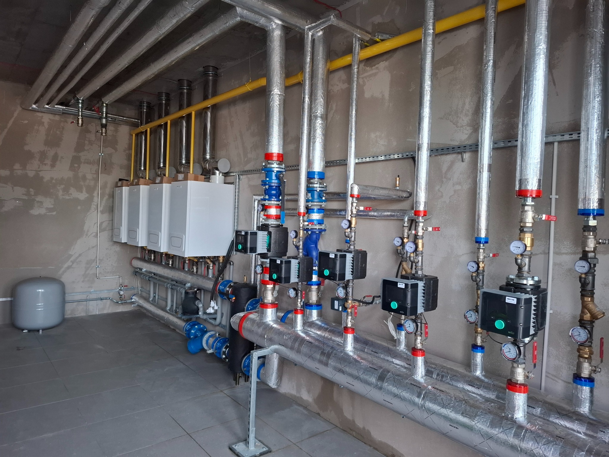 Kompleksowa instalacja grzewcza: kotły, rury w izolacji, zawory, manometry. Widoczne elementy instalacji gazowej i grzewczej w surowym wnętrzu.