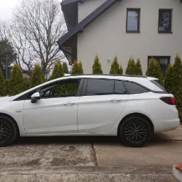 Białe kombi z przyciemnionymi szybami bocznymi i tylną, zaparkowane przed domem z tujami w tle.