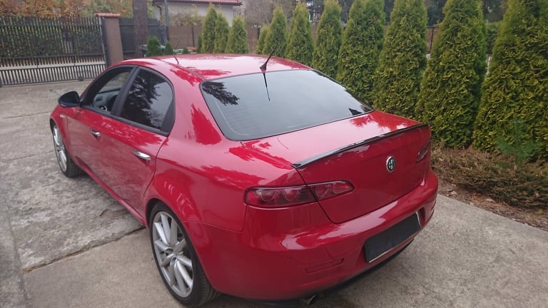 Czerwona Alfa Romeo 159 z przyciemnionymi szybami, widziana z tyłu na betonowym podjeździe, z żywopłotem z tui w tle.
