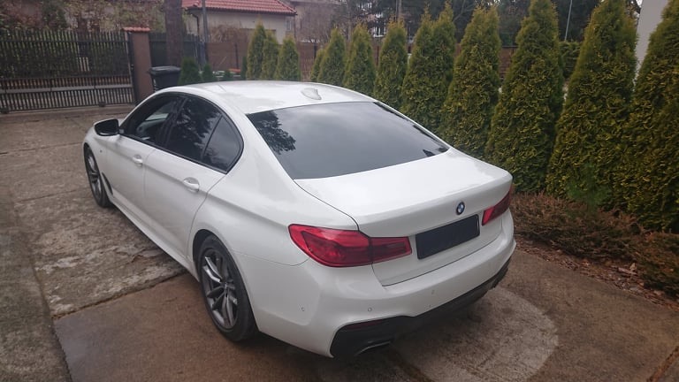 Białe BMW serii 5 z przyciemnionymi szybami tylnymi, zaparkowane na betonowym podjeździe przed domem z tujami w tle.