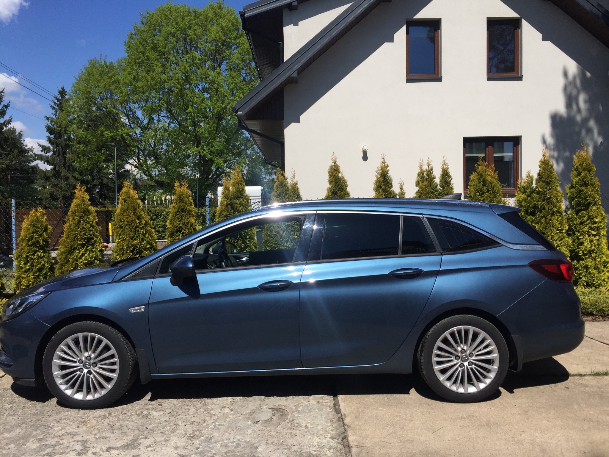 Niebieska Opel Astra kombi z przyciemnionymi szybami, zaparkowana przed domem w Warszawie w słoneczny dzień.