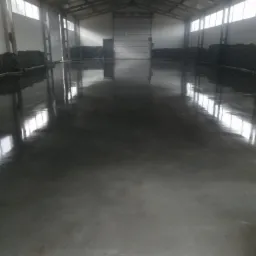 Świeżo wylana, ciemnoszara posadzka w hali przemysłowej, odbijająca światło z okien i bramy wjazdowej.