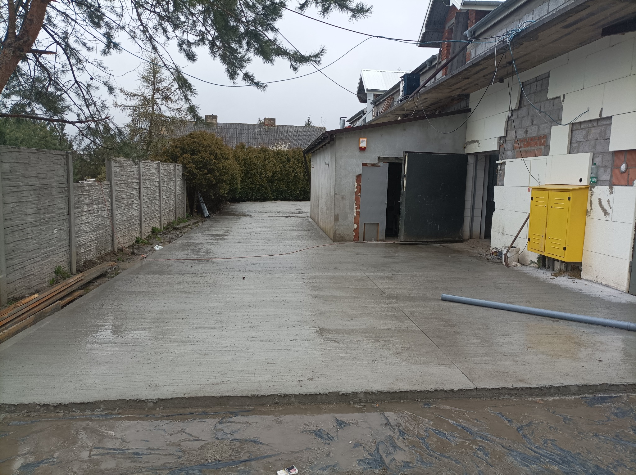 Świeżo wylana, gładka posadzka przemysłowa na zewnątrz budynku, z widocznymi szczelinami dylatacyjnymi i szarym rurociągiem leżącym na powierzchni. Budynek w trakcie ocieplania styropianem, żółta...