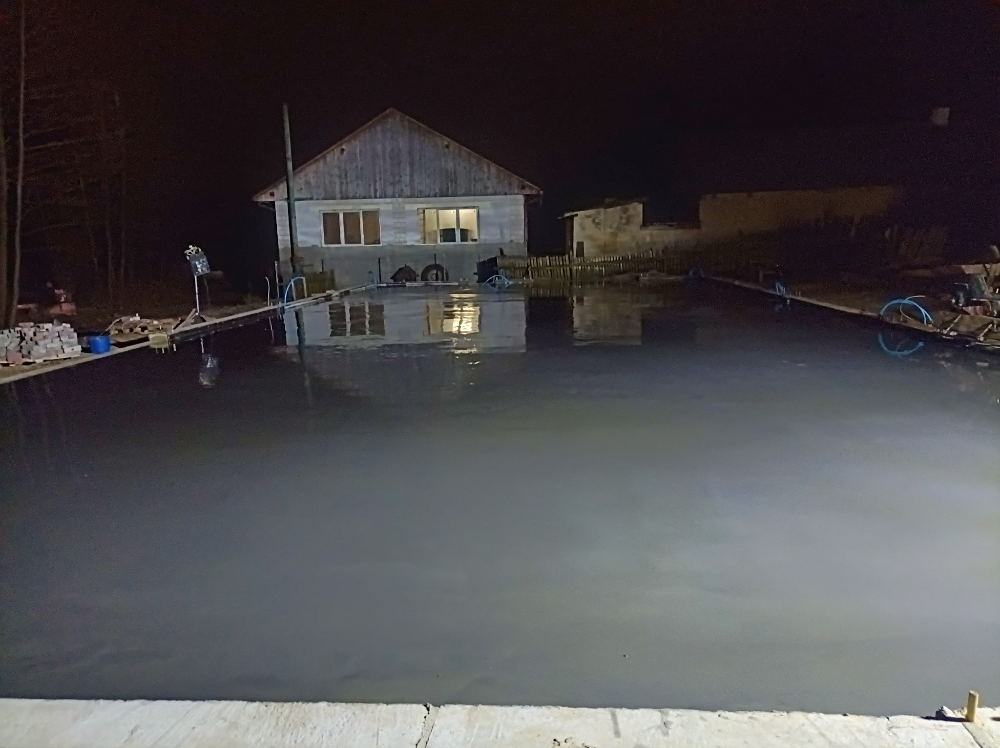 Świeżo wylana, gładka, szara wylewka pod posadzkę przemysłową w słabym oświetleniu nocnym, z widocznym budynkiem w tle i sprzętem budowlanym po bokach.