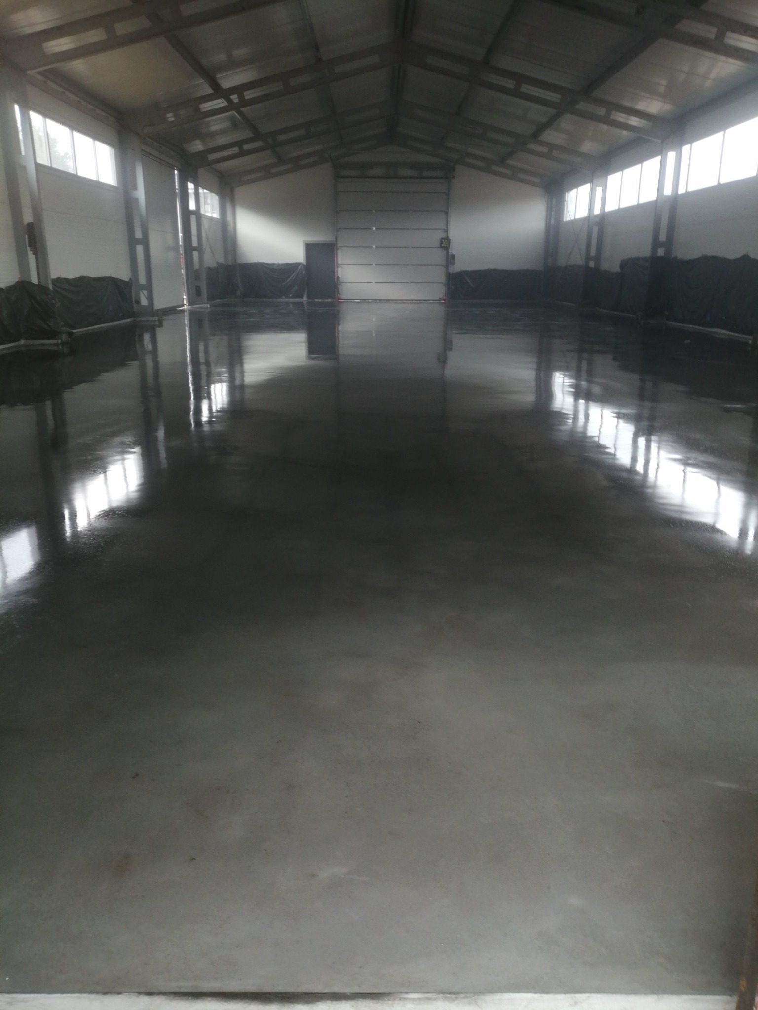 Świeżo wylana, ciemnoszara posadzka w hali przemysłowej, odbijająca światło z okien i bramy wjazdowej.