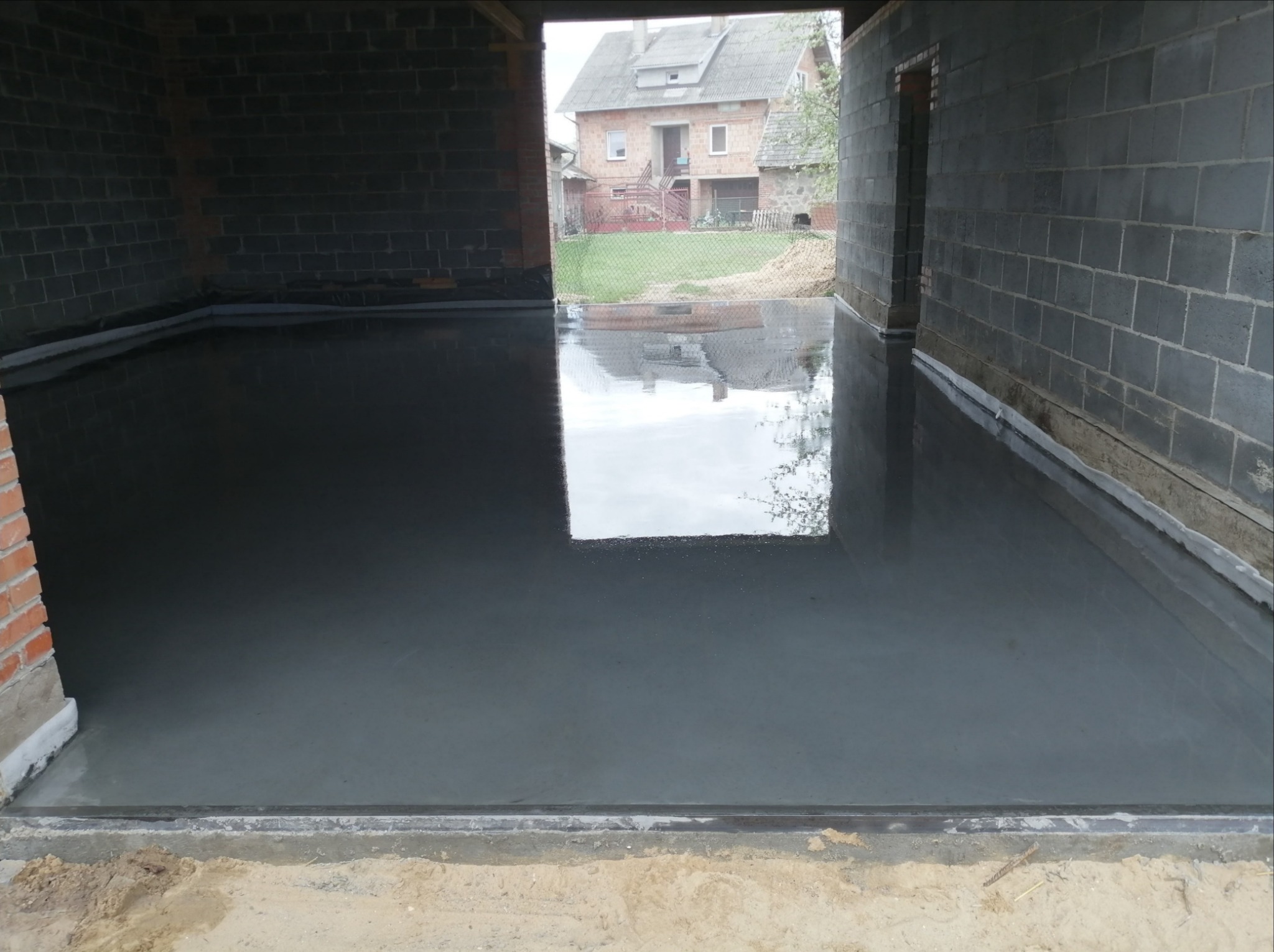 Świeżo wylana, gładka posadzka przemysłowa w surowym garażu z bloków betonowych, z widocznym odbiciem budynku na zewnątrz.