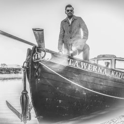 Igor Skarżyński - Fotografia - Czarno-białe zdjęcie mężczyzny w płaszczu i okularach przeciwsłonecznych, stojącego na dziobie łodzi z napisem 'TAWERNA KAPITAŃSKA' w Lublinie, z widocznym kotwicą i rzeką w tle.