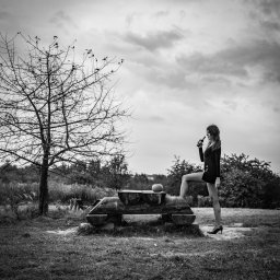Igor Skarżyński - Fotografia - Czarno-białe zdjęcie kobiety w czarnym ubraniu stojącej z jedną nogą na drewnianej konstrukcji, trzymającej butelkę w plenerze z drzewem bez liści i pochmurnym niebem w tle.