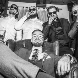 Igor Skarżyński - Fotografia - Czarno-białe zdjęcie grupy eleganckich mężczyzn z cygarami i drinkiem, siedzących i stojących w pomieszczeniu z drewnianym sufitem. Jeden mężczyzna siedzi w fotelu.