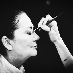 Igor Skarżyński - Fotografia - Czarno-białe zbliżenie profilu kobiety podczas makijażu oka pędzlem. Delikatny pierścionek na palcu, minimalistyczna bransoletka na nadgarstku, ciemne tło.