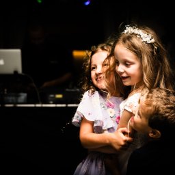 Igor Skarżyński - Fotografia - Trójka dzieci w objęciach na tle ciemnego pomieszczenia z laptopem. Dziewczynka w wianku, uśmiechnięta, w jasnej sukience. Ujęcie z dołu, oświetlenie punktowe.