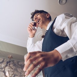 Igor Skarżyński - Fotografia - Uśmiechnięty mężczyzna w kamizelce pije z kieliszka, trzymając pustą szklankę. Nietypowa perspektywa z dołu, widoczne dekoracje na ścianie.