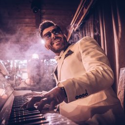 Igor Skarżyński - Fotografia - Uśmiechnięty mężczyzna w białym smokingu i okularach gra na białym fortepianie w zadymionym pomieszczeniu. Widoczne ręce na klawiszach i fragment dekoracji.