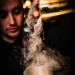 Igor Skarżyński - Fotografia - Barman z brodą nalewa dymiący koktajl z złotego shakera do szklanki. Efektowny drink w ciemnym otoczeniu, z rozmytymi światłami w tle.