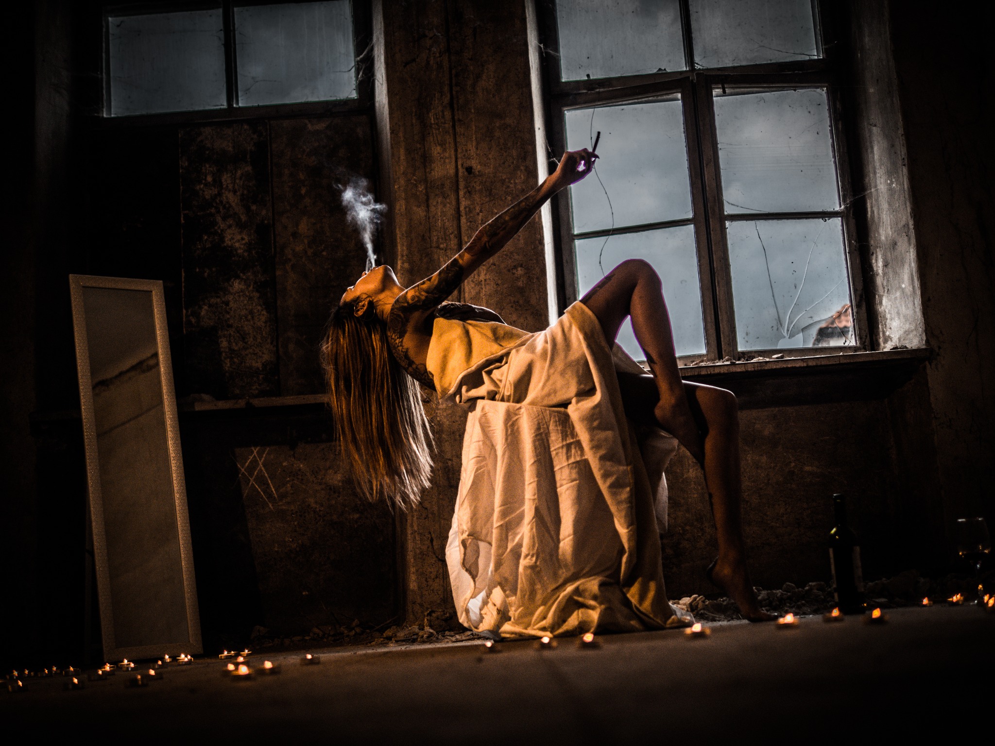Kobieta z tatuażami, leżąca na podwyższeniu, owinięta w jasną tkaninę, pali papierosa w opuszczonym pomieszczeniu z oknami z powybijanymi szybami, oświetlonym świecami.
