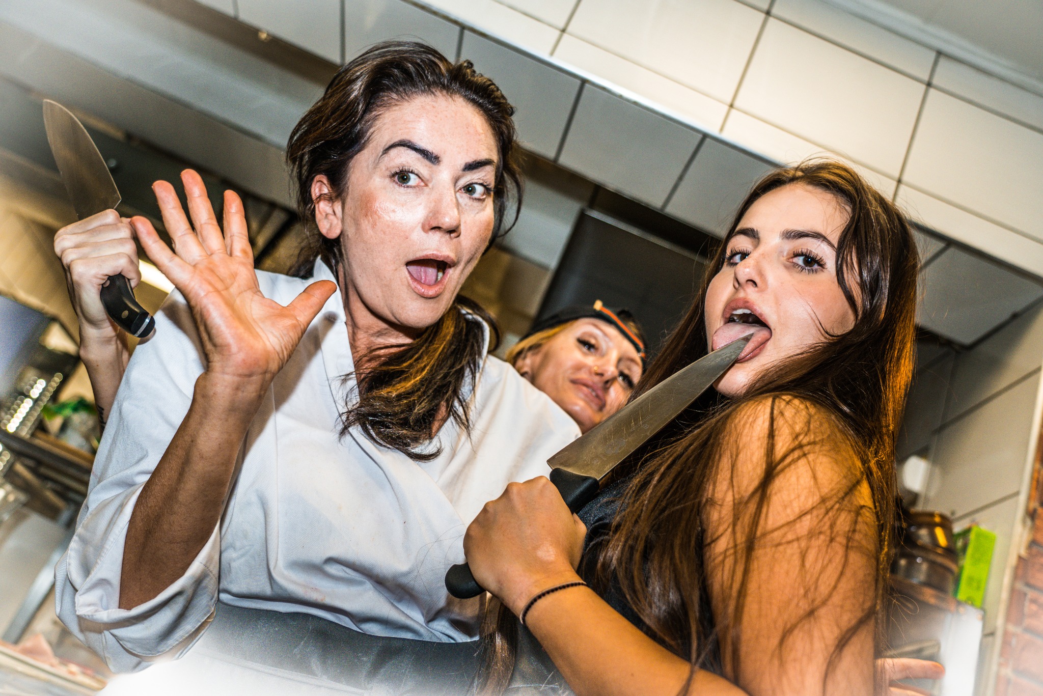 Kobieta w białej bluzie z otwartymi ustami i uniesioną ręką, a obok niej dziewczyna z wyciągniętym językiem dotykającym ostrza noża, w tle trzecia kobieta.