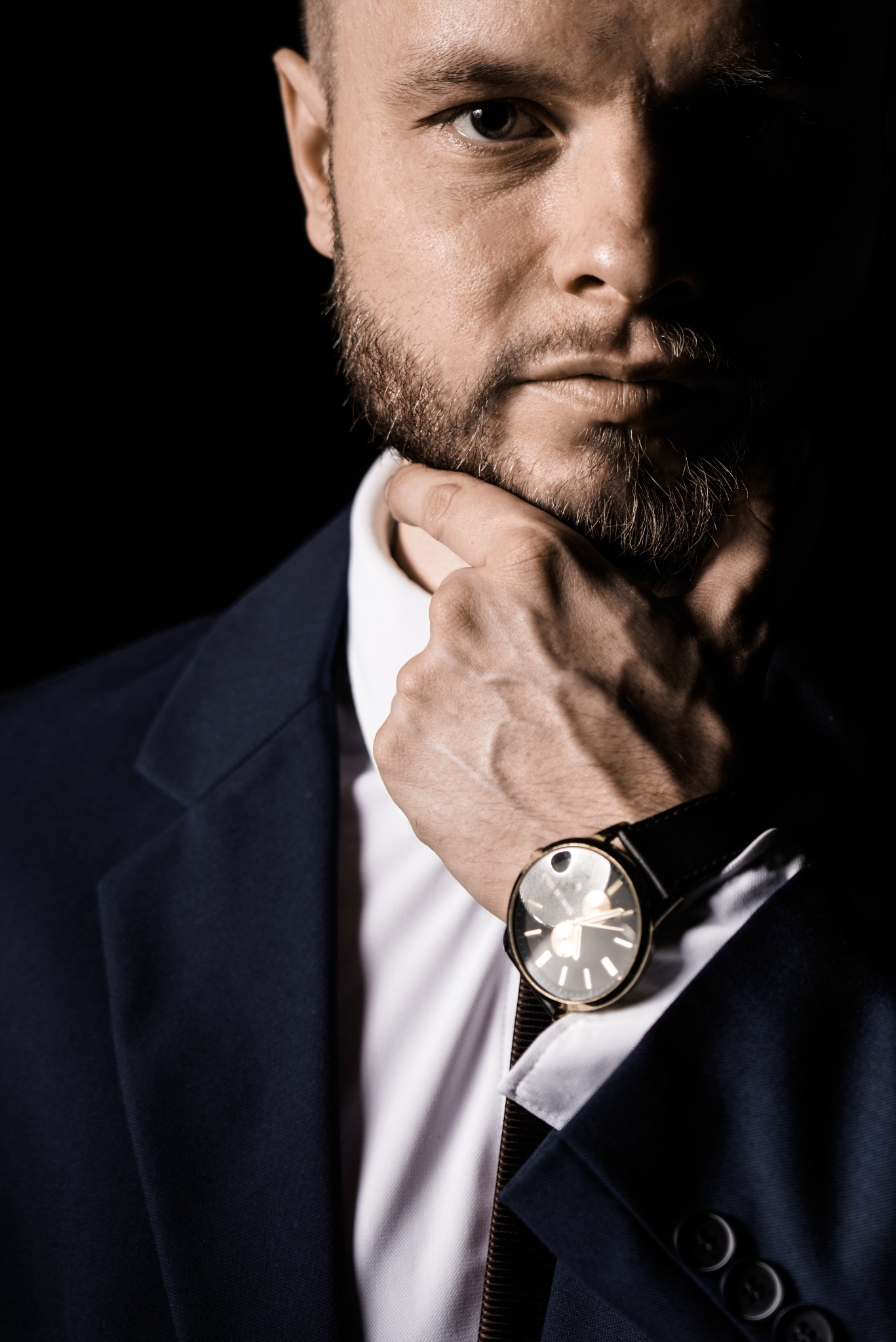 Portret mężczyzny z brodą w garniturze, z zegarkiem na ręce, w dramatycznym oświetleniu, dotykającego brody palcem.