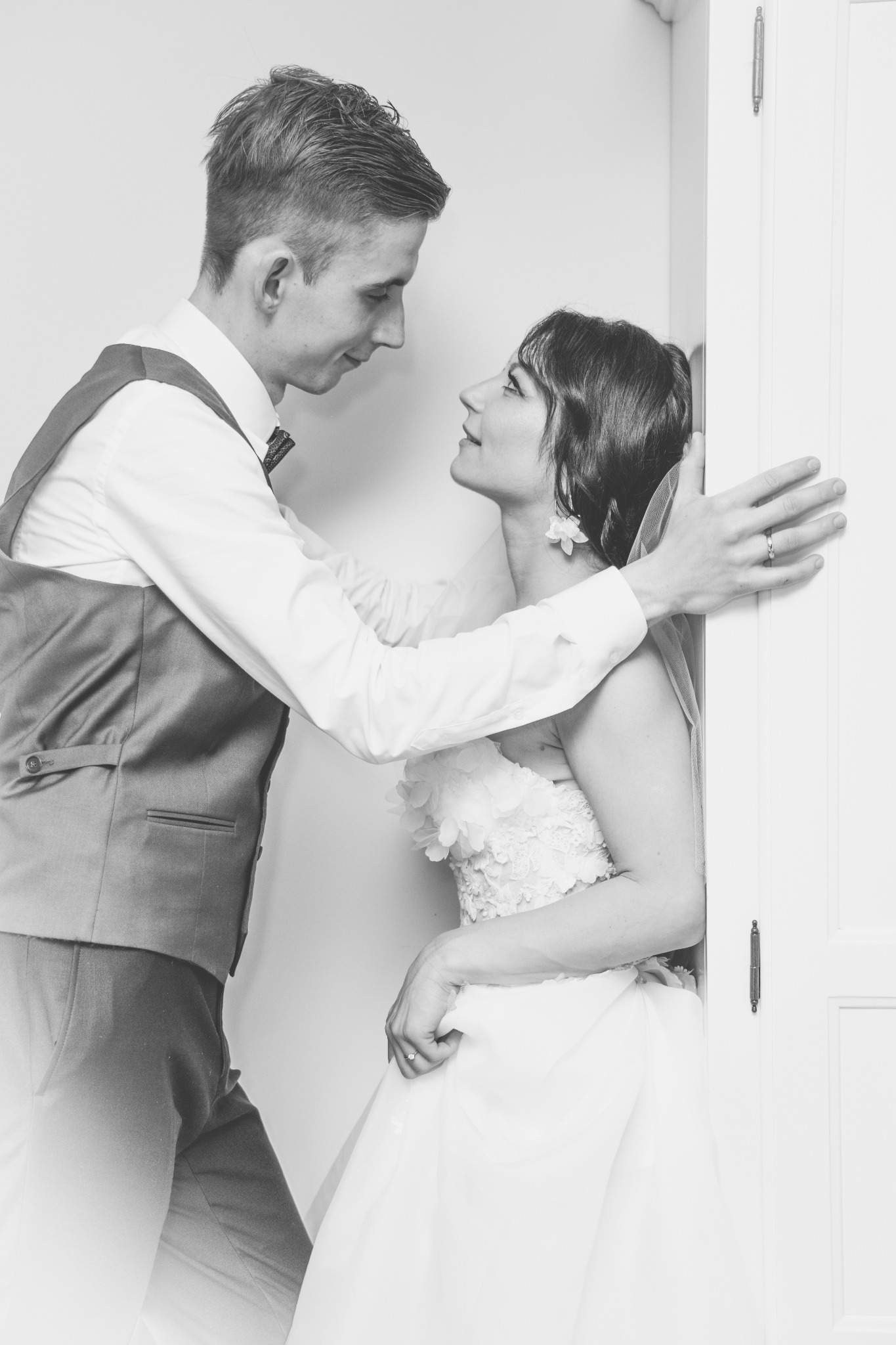 Czarno-białe zdjęcie pary młodej: Panna młoda w sukni z kwiatowymi aplikacjami i welonie, oparta o białe drzwi, spogląda z uśmiechem na Pana młodego w kamizelce i muszce.