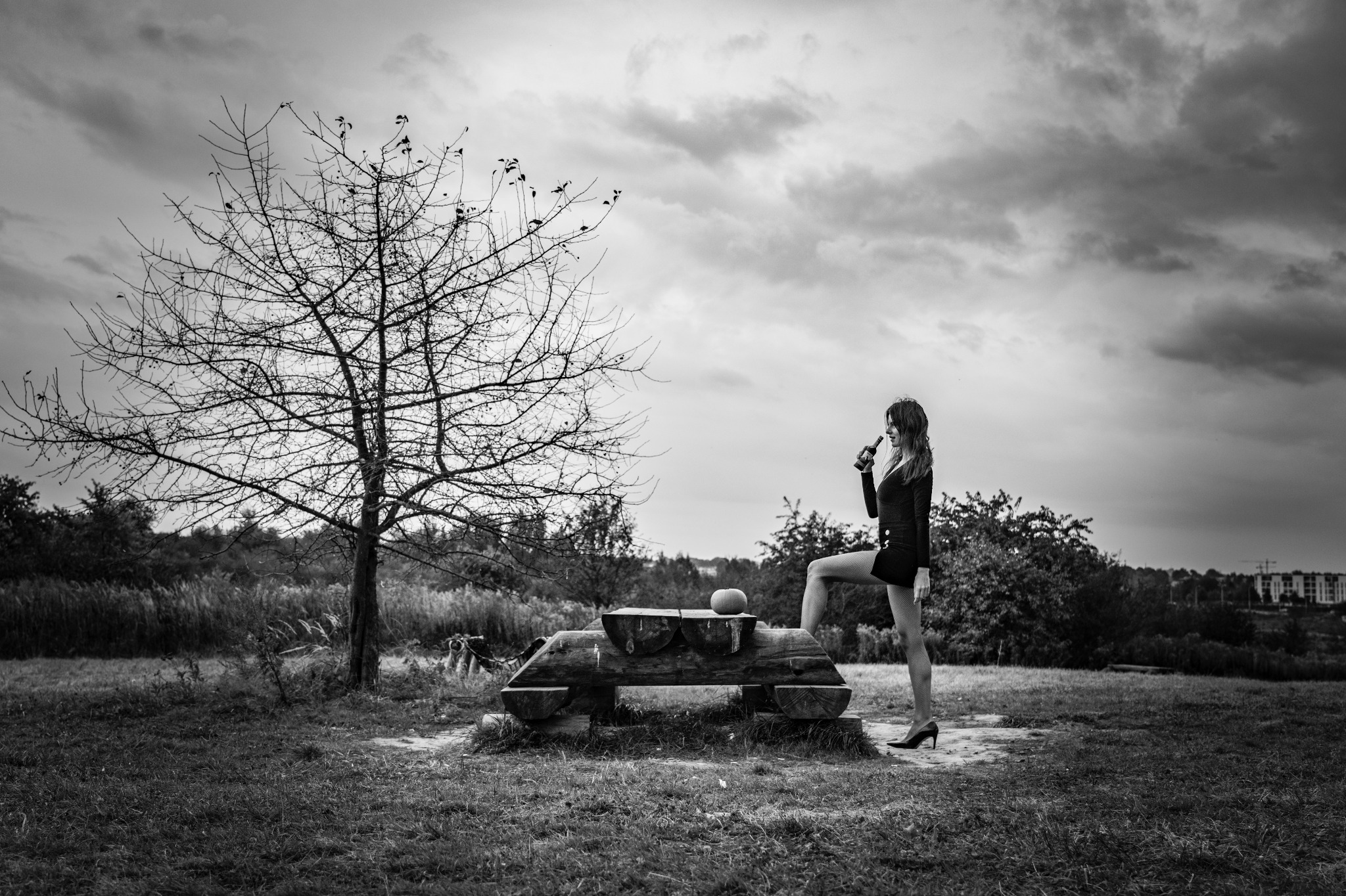 Czarno-białe zdjęcie kobiety w czarnym ubraniu stojącej z jedną nogą na drewnianej konstrukcji, trzymającej butelkę w plenerze z drzewem bez liści i pochmurnym niebem w tle.