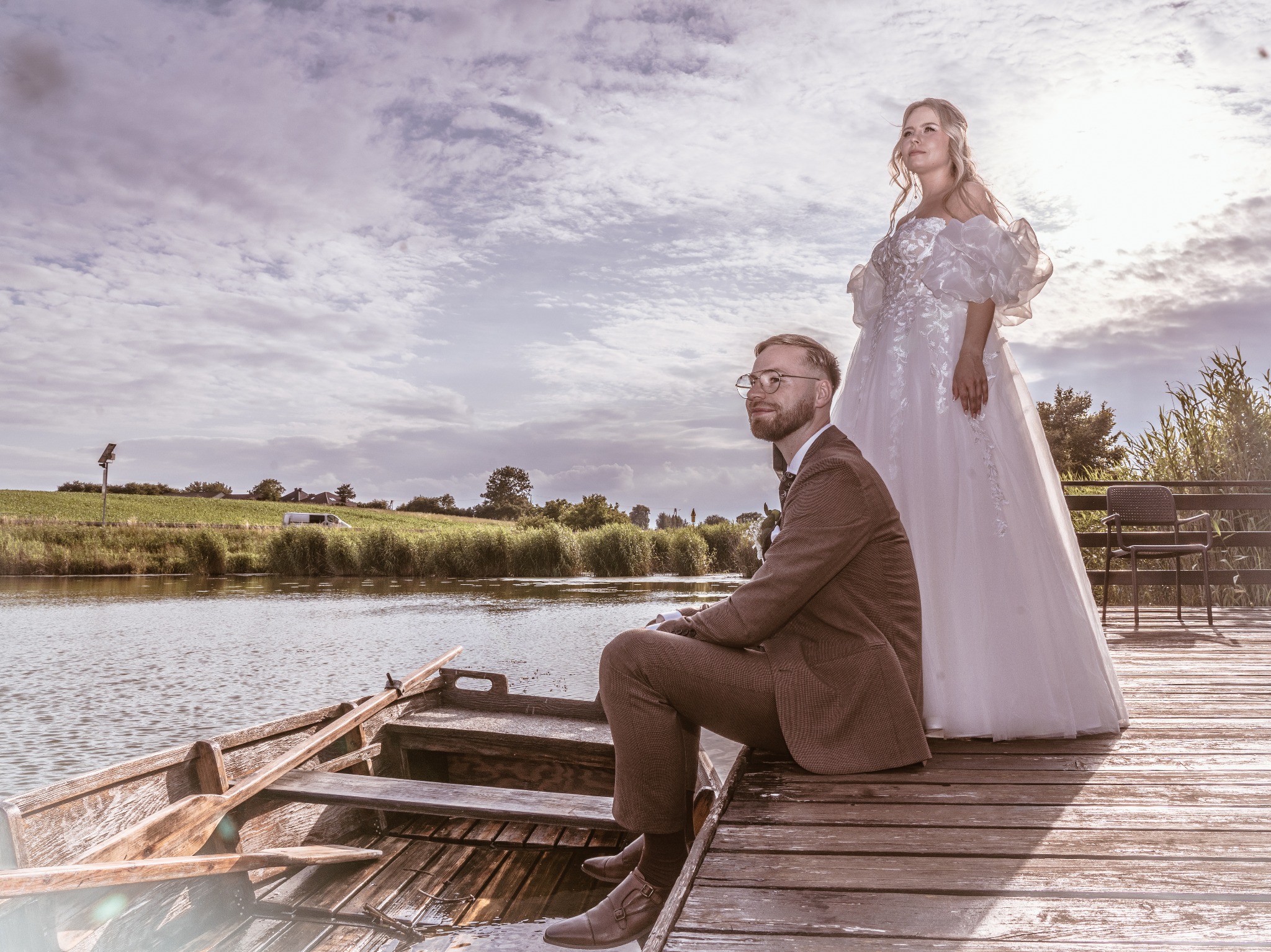 Elegancka sesja narzeczeńska: para w łodzi na jeziorze, on w brązowym garniturze, ona w białej sukni ślubnej, na tle zachmurzonego nieba i zieleni.