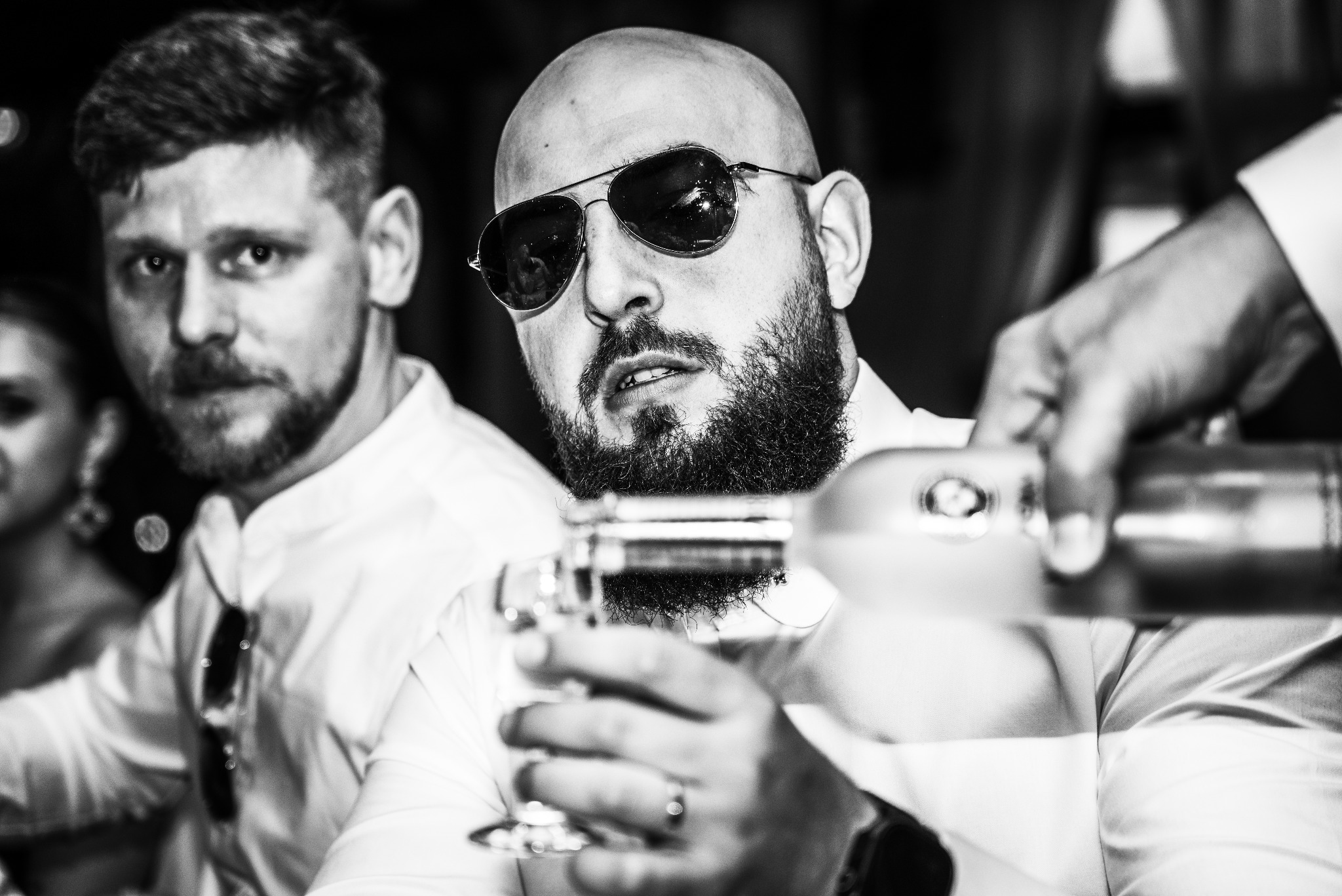 Czarno-białe ujęcie mężczyzny w okularach nalewającego alkohol do kieliszka, drugi mężczyzna w tle, elegancka atmosfera.