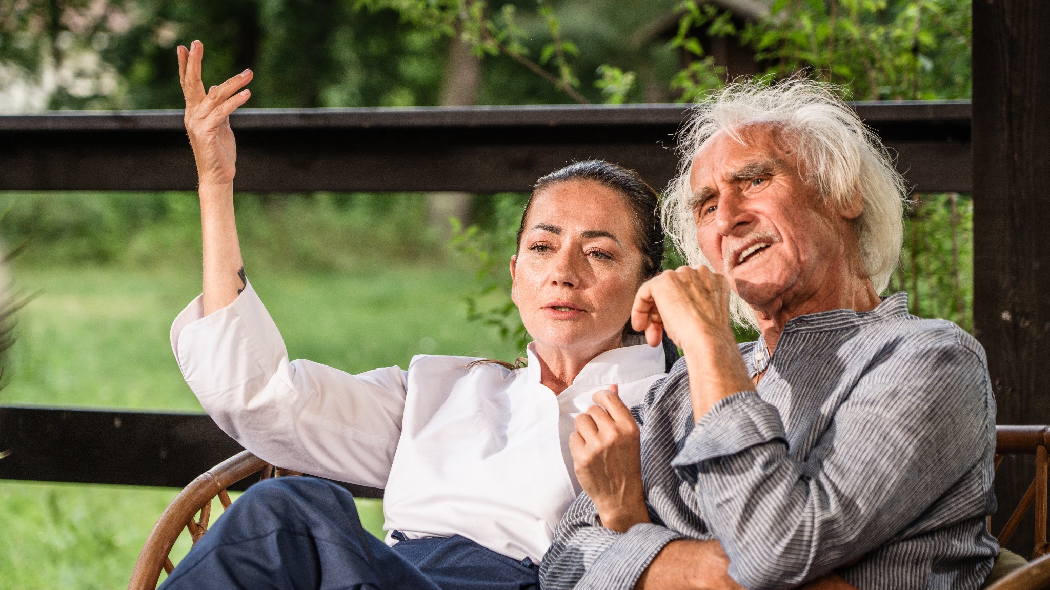 Para w średnim wieku siedzi w wiklinowym fotelu na tle zieleni. Kobieta gestykuluje ręką, mężczyzna patrzy w dal. Ubrani w jasne koszule, swobodna atmosfera.