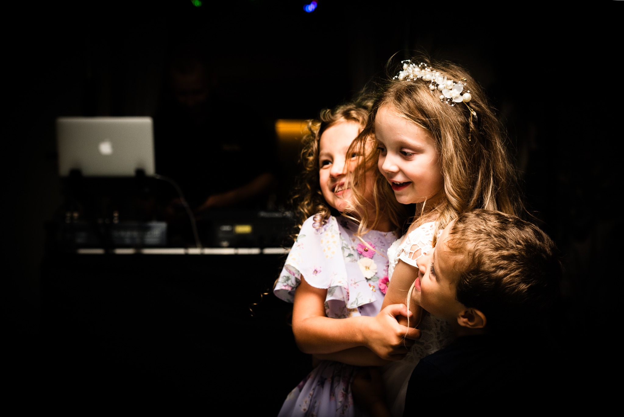 Trójka dzieci w objęciach na tle ciemnego pomieszczenia z laptopem. Dziewczynka w wianku, uśmiechnięta, w jasnej sukience. Ujęcie z dołu, oświetlenie punktowe.