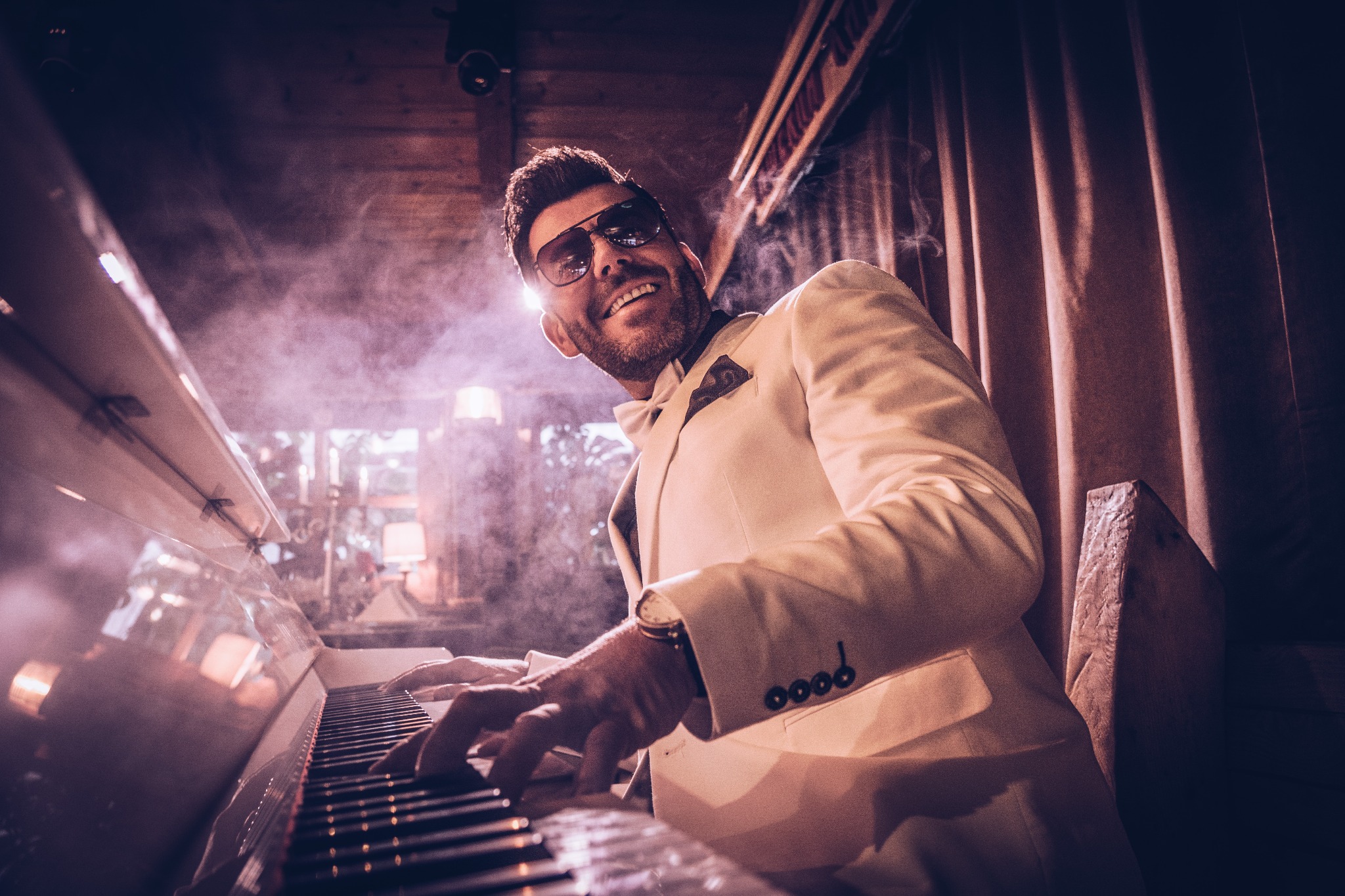 Uśmiechnięty mężczyzna w białym smokingu i okularach gra na białym fortepianie w zadymionym pomieszczeniu. Widoczne ręce na klawiszach i fragment dekoracji.