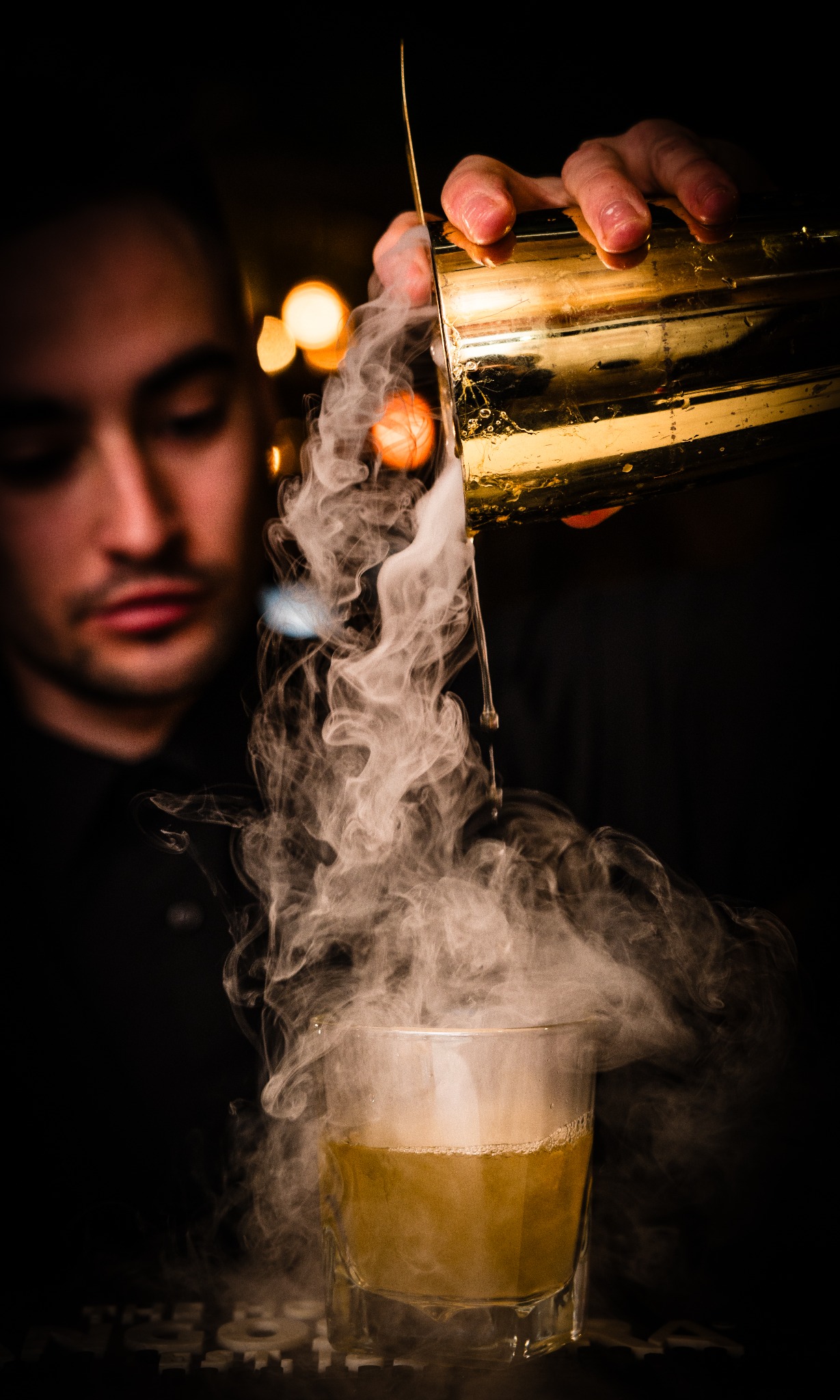 Barman z brodą nalewa dymiący koktajl z złotego shakera do szklanki. Efektowny drink w ciemnym otoczeniu, z rozmytymi światłami w tle.