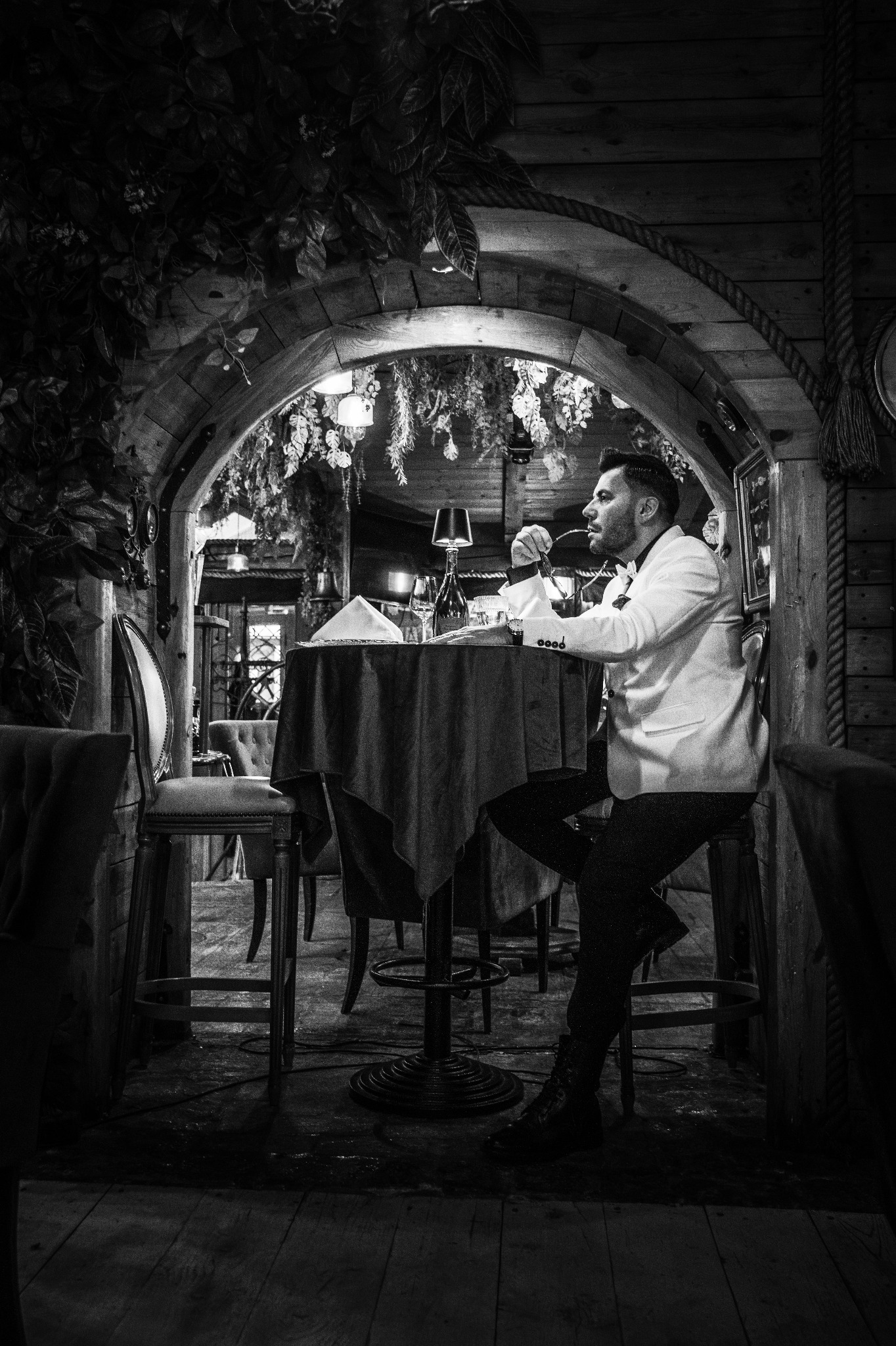 Czarno-białe zdjęcie mężczyzny w białej marynarce siedzącego przy stole w restauracji o rustykalnym wystroju, z dekoracjami roślinnymi i drewnianymi elementami.