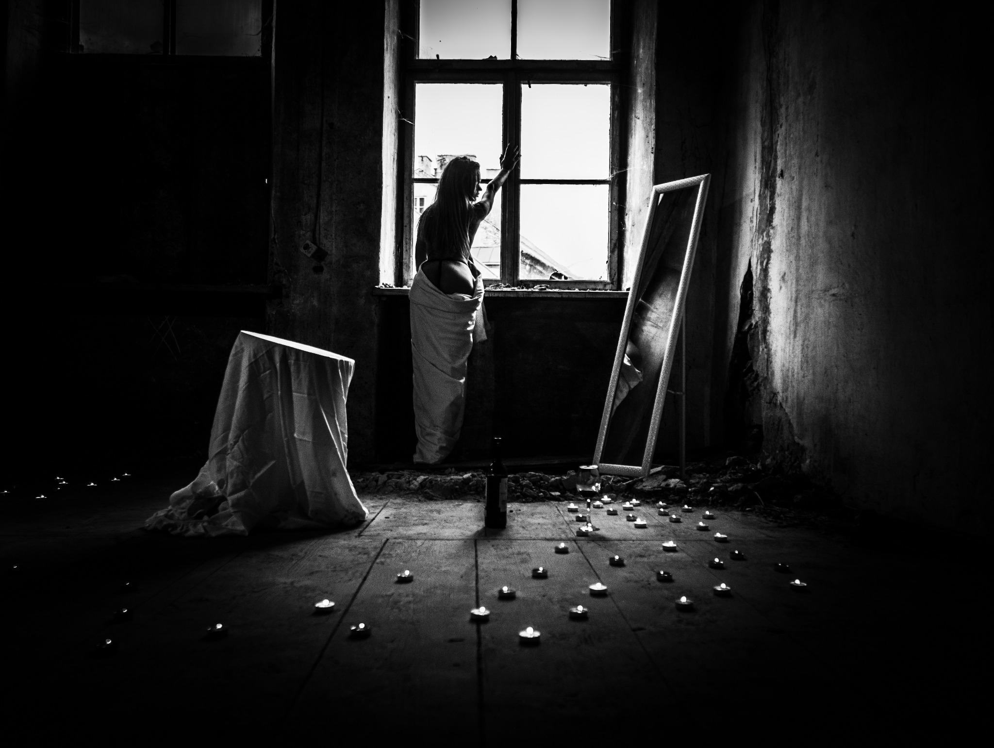 Czarno-białe zdjęcie kobiety w oknie, owiniętej w materiał, otoczonej świecami, z lustrem w tle, w surowym wnętrzu. Klimatyczne ujęcie z artystycznym zamysłem.