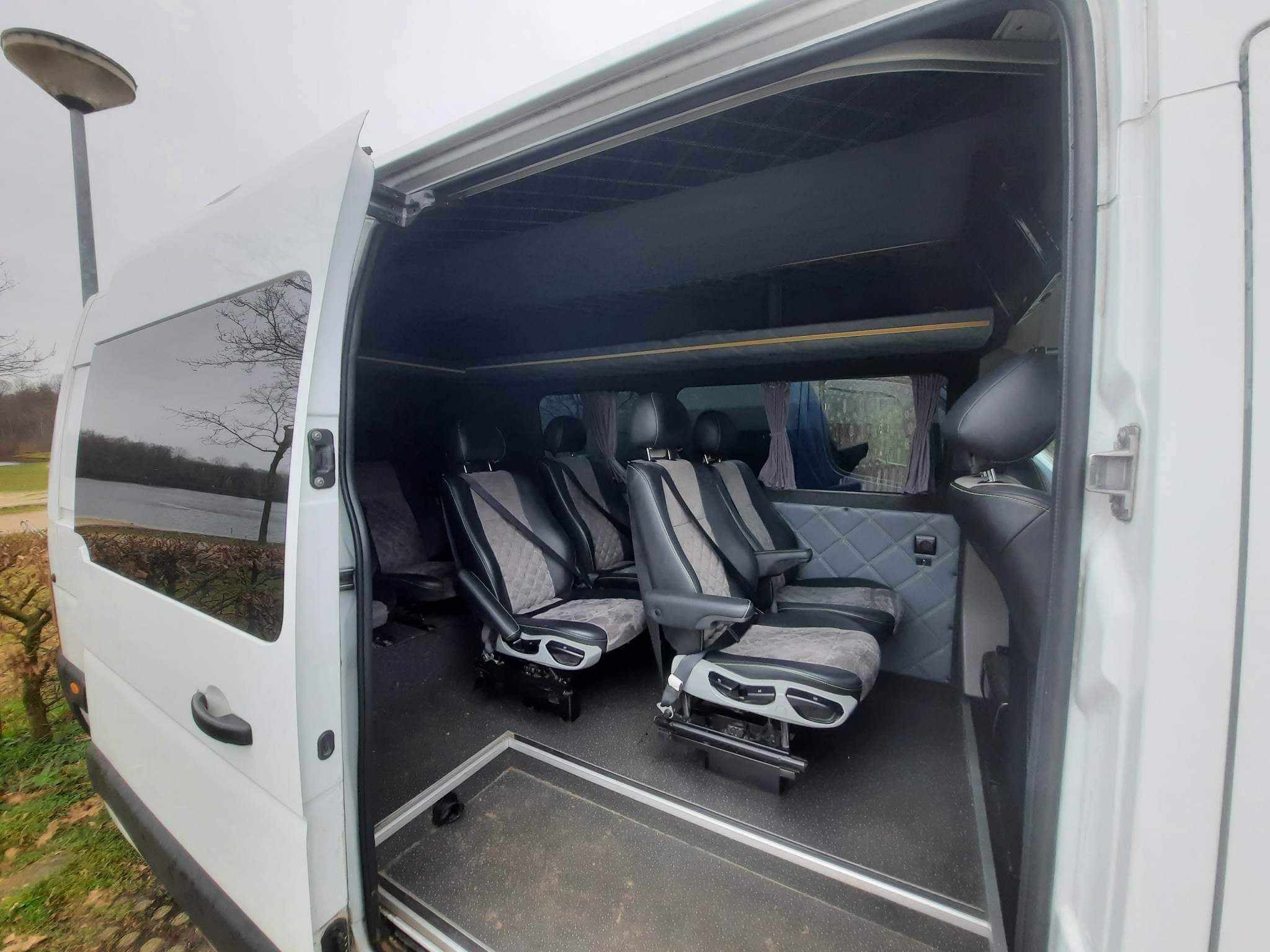 Wnętrze białego busa z komfortowymi, szaro-czarnymi fotelami z podłokietnikami i przeszyciami w romby, widok z otwartych drzwi bocznych na szary, wyściełany sufit i zasłonki w oknach.