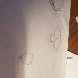 Szkic oł&oacute;wkiem na papierze przedstawiający r&oacute;żne projekty pierścionk&oacute;w, w tym jeden z motywem serca i otwarte pudełko na pierścionek.